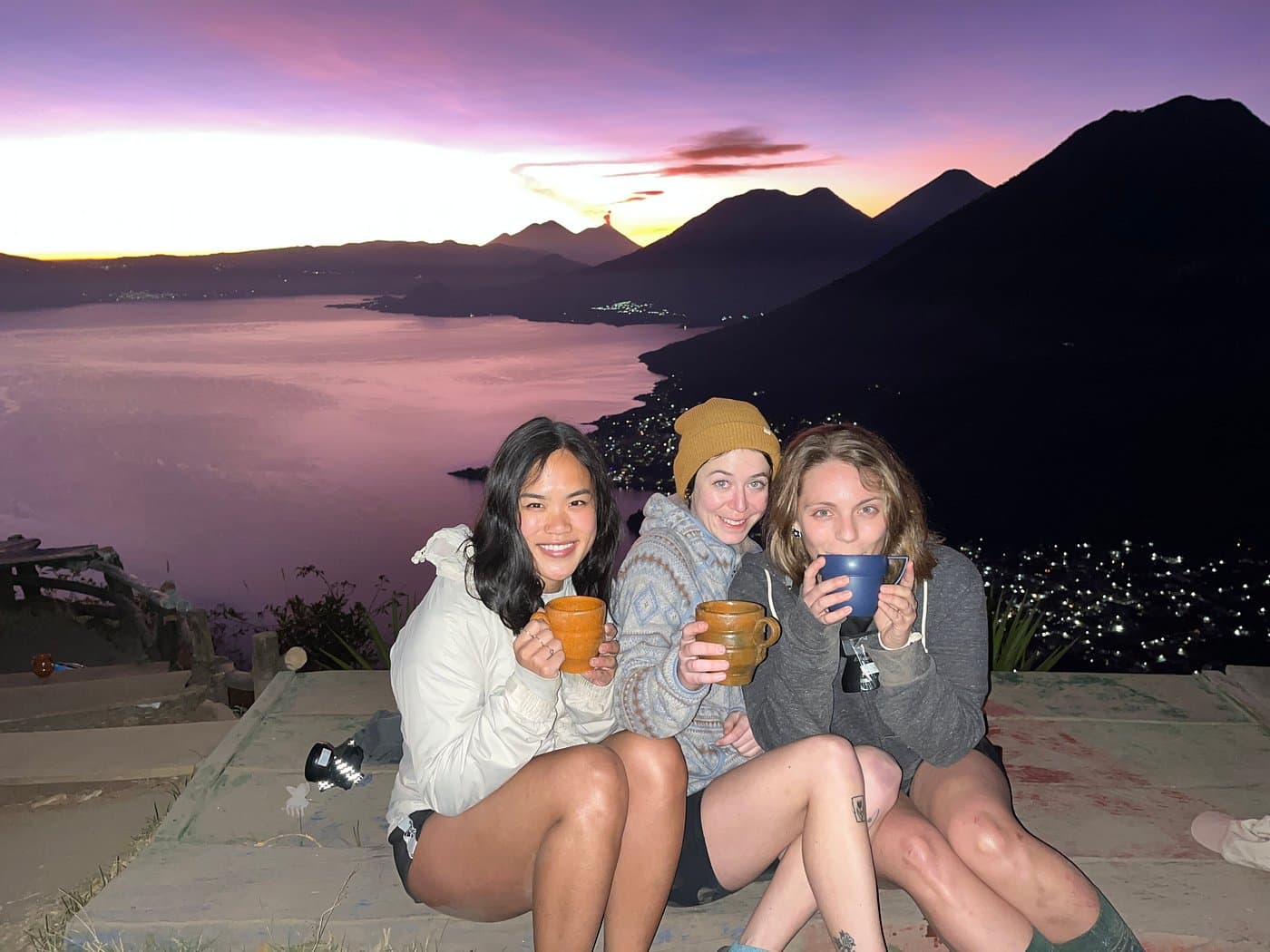 Three hikers with mugs of hot chocolate at the summit, the Fuego volcano erupting on the horizon behind Lake Atitlán